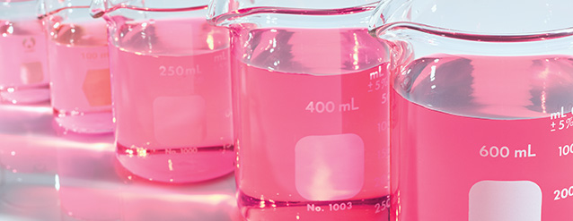 Mary Kay beakers holding pink, orange and red liquids standing against a white background.