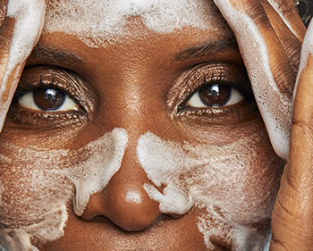 Знімок зблизька, на якому зріла жінка вмиває обличчя руками, на щоках, носі та лобі є біла мильна піна