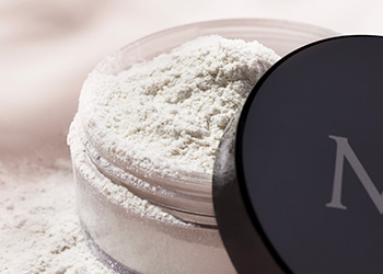 Up-close shot of opened jar of Mary Kay® Translucent Loose Powder with lid resting on base of package and loose powder spilling over the edge over a solid background with shadow overlays