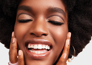 Smiling black model with curly afro hairstyle showcasing cut crease eyeshadow look, hands resting on cheeks, looking confident and stylish.