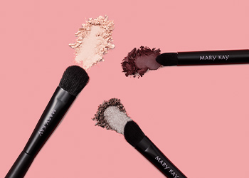 3 Mary Kay Eye Shadow brushes each with eye shadow rub behind them in different shades from light to dark neutrals on a pink background.