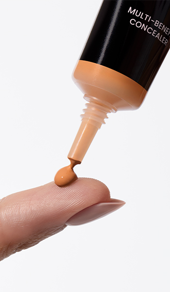 Відкритий тюбик Коректору для шкіри навколо очей Mary Kay сфотографований разом із аплікатором та пензликом для макіяжу кремового кольору на рожевому тлі із мазком засобу.