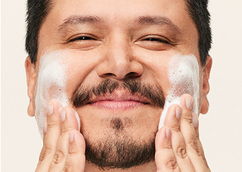 A dark-haired man with facial hair happily washing his face