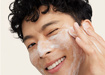 A young, curly-haired man happily cleansing his skin and winking at the camera